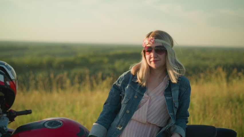 A woman wearing a denim jacket and pink gown smiles while resting on a motorcycle in an open field, she wears sunglasses, giving off a relaxed and carefree vibe