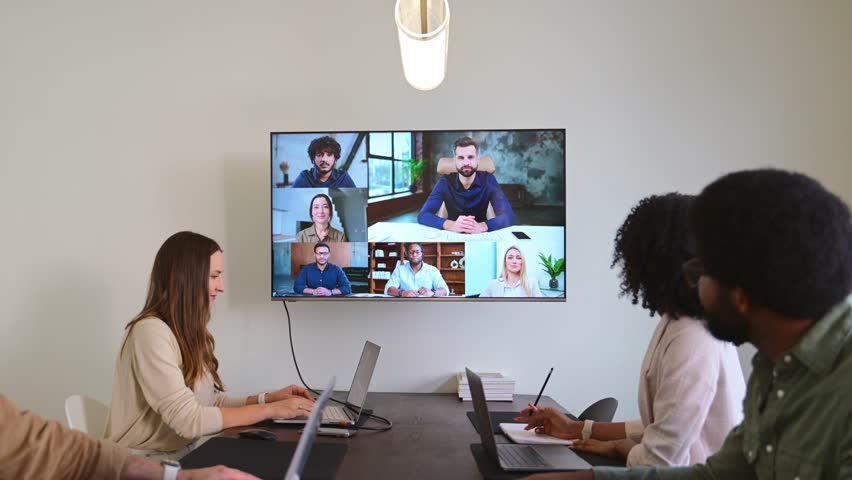 A hybrid team meeting where in-person attendees discuss and collaborate with remote colleagues through a video conference displayed on a wall-mounted screen. Concept of connectivity and flexibility - Powered by Shutterstock - Get 15% off with code: PIKWIZARD15