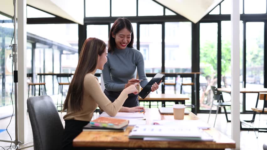 Two young Asian businesswoman working on digital tablet with analysis report and advisor showing plan, Financial analysis and Marketing teamwork concept.