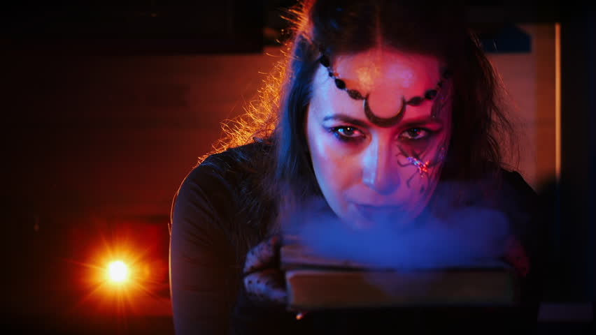 Close-up of a girl dressed as a witch, looking at the camera and holding a smoking old book. Horror and Halloween concept.