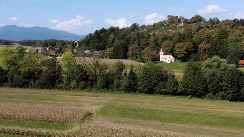  A peaceful countryside scene with a small white church on a hill, surrounded by trees and fields of corn. Scattered houses rest on a distant hill under a blue sky with clouds.