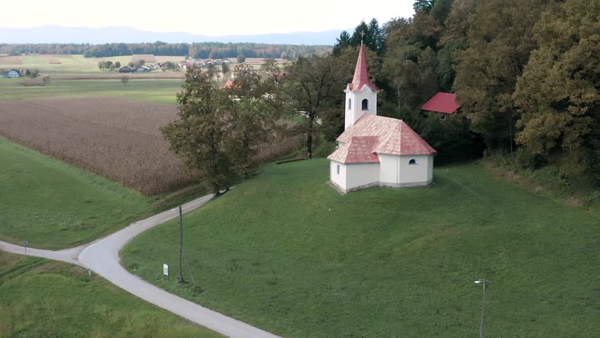 A quaint white church with a pointed red roof stands atop a green hill, framed by a winding path that cuts through farmlands. Cornfields stretch across the foreground, while trees surround the church.
