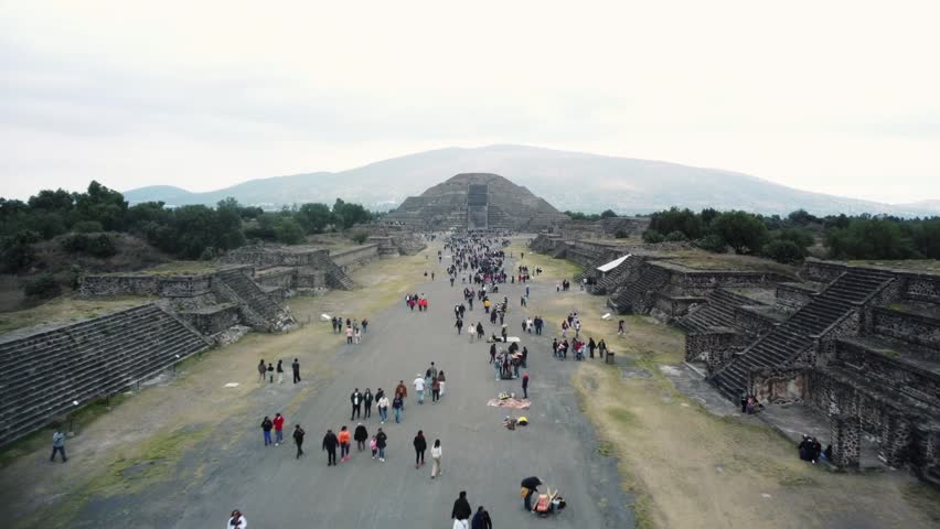 Aerial footage of the Pyramid of the Moon and the Avenue of the Dead in Teotihuacan, Mexico. Tourists walk through the historic site, offering a stunning view of ancient pyramids and plazas.