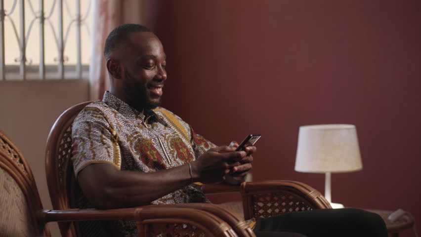 A young African man smiles whiles looking at his phone. He is doing some banking transactions at home on his smartphone.