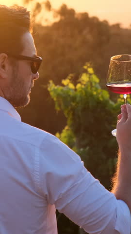 young man tasting red wine in a vineyard during sunset. stylish man in white shirt holding glass of red wine and enjoying sunset in vineyard. Close up of sommelier man sniffing wine in glass vertical