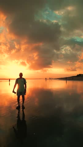 A person stands on a beach in Bali during a vivid sunset, with the sky reflecting on the wet sand.