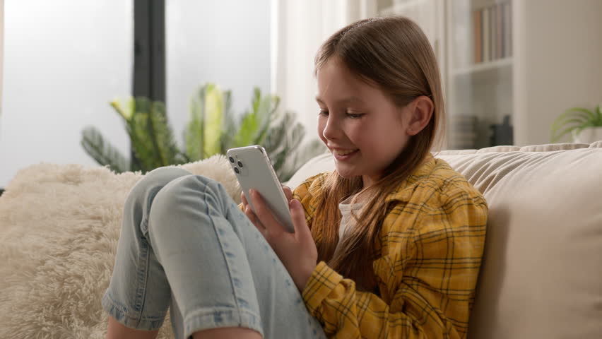 Gadget addiction. Cheerful positive Caucasian child preteen girl daughter smile typing mobile phone device scrolling feed social media little kid playing virtual games chatting online sofa home alone