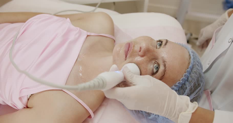 A young woman in a pink gown lies comfortably during a facial treatment in a modern spa, attended by a healthcare professional. Woman Receives Professional Facial Treatment in Spa Salon