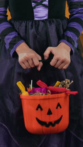 Medium vertical shot of hands and midsection of unrecognizable young girl in black and purple witch dress posing on orange background in studio with pumpkin bucket full of Halloween sweets