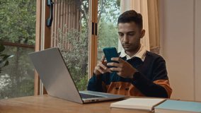 Medium shot of young Caucasian man using wired headphones and smartphone while working at home desk with laptop - Powered by Shutterstock - Get 15% off with code: PIKWIZARD15