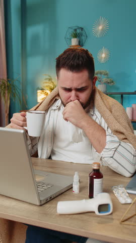 Sick ill young businessman suffering from cold flu or allergy working on laptop at home office. Manager freelancer man wrapped in blanket blows sneezes wipes snot into napkin. Coronavirus quarantine.