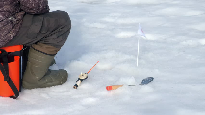 Ice fisherman sitting patiently with rod and flag indicator on fishing competition. Man catching fish on frozen river with ice and snow. Outdoor winter activity hobby leisure recreation kind of sport.
