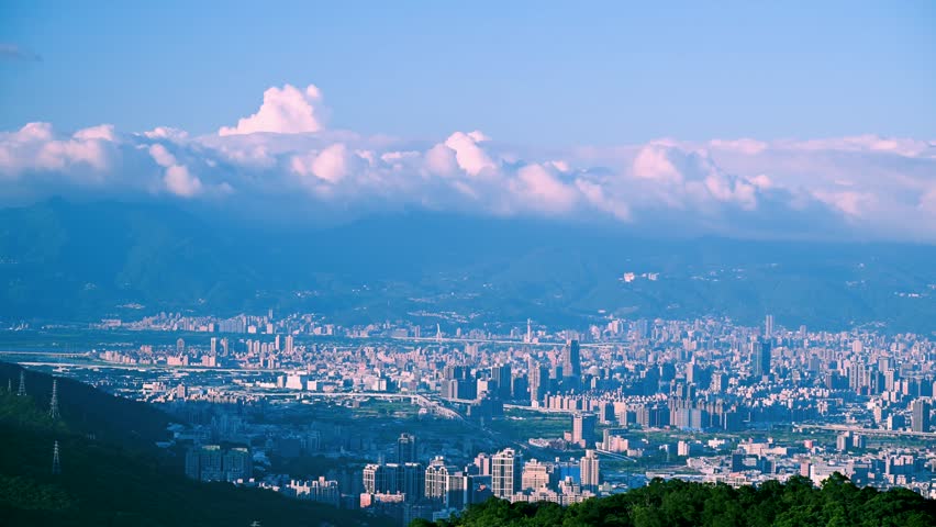 A breathtaking panoramic view of a bustling city at sunset. Soft, fluffy clouds drift across the sky, casting a warm glow over the urban landscape. Captured in Dading Mountain, Shulin, Taiwan.