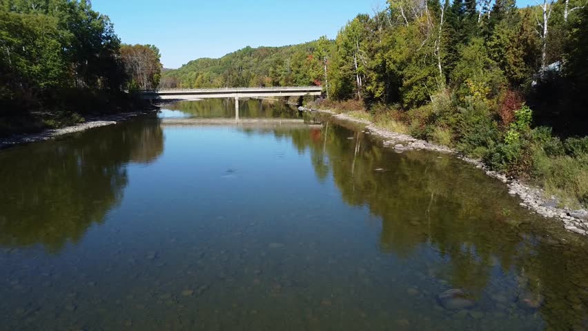 Aerial approach at low altitude to a concrete bridge spanning a river with calm and clear water reflecting the colors of the surrounding scenery.  Matane Salmon River, Quebec, Canada, 2025.