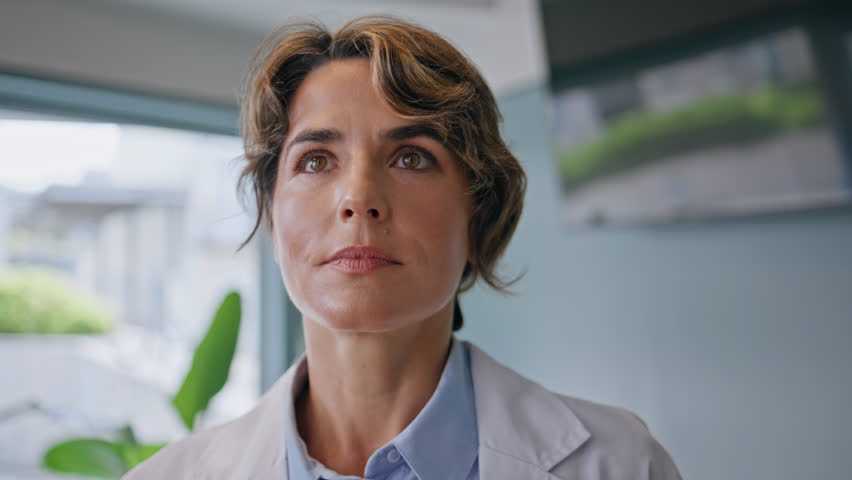 Portrait of calm woman physician standing at healthcare office hall. Serious professional doctor looking camera wearing white robe closeup. Experienced general practitioner posing in modern clinic.