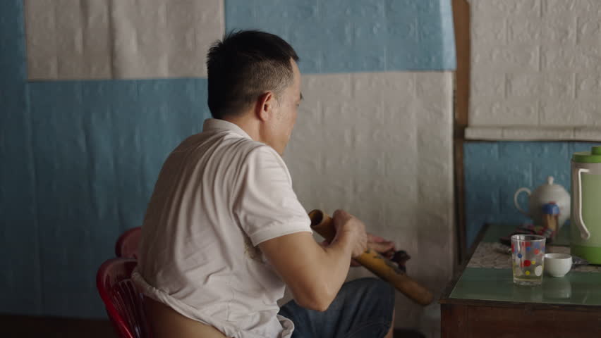 mature asian man drinks soda and smokes tobacco from a large handmade wooden pipe in vietnam