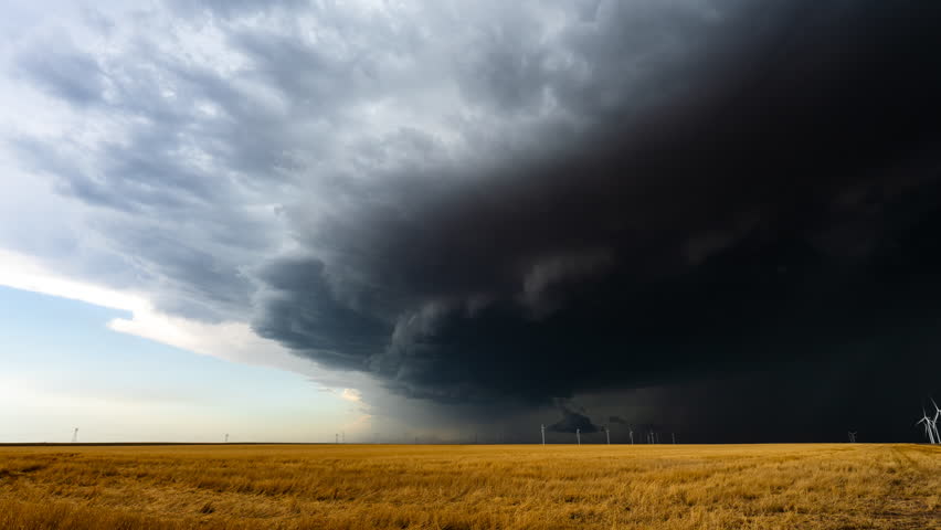 Storm clouds roll across the sky, casting shadows over warm-colored fields and a wind farm, creating a striking contrast of nature and technology.