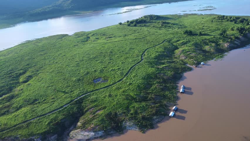 Aerial view of dirt road in green field under cloudy sky in rainy season by lake in rural.