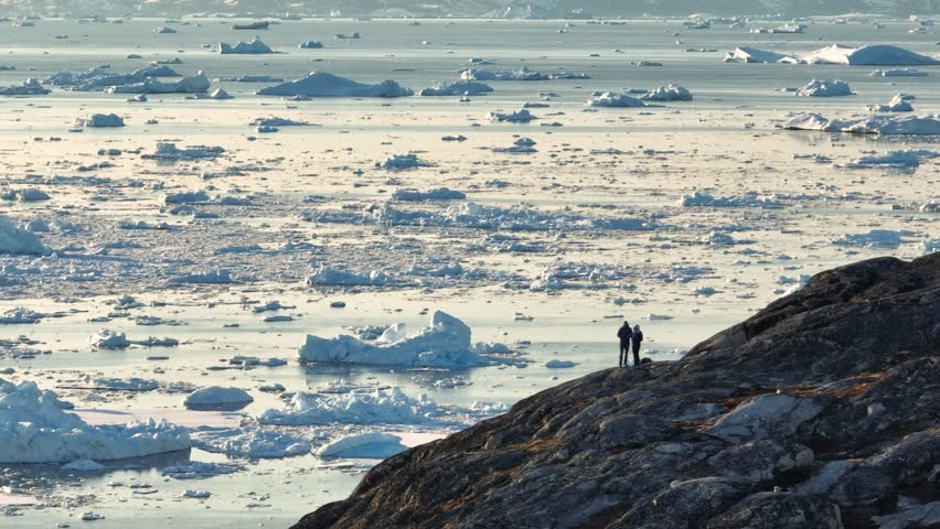 Two people on cliff watch melting ice in the Pacific Ocean. Aerial shot of couple travels to Greenland. Icebergs in the ocean near Ilulissat, Greenland