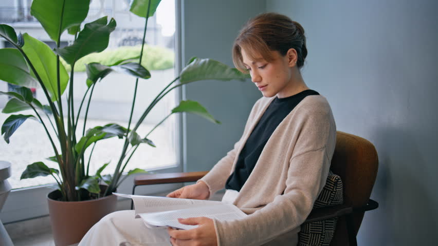 Woman visitor reading calmly in waiting room of healthcare clinic closeup. Relaxed patient sitting by hall window focused on magazine. Attractive lady relaxing before meeting doctor in medical office.