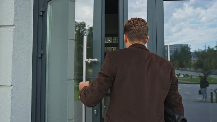 A professional man walks through a glass door into a sleek office building. The scene showcases a bright day, reflecting urban life and business activities in the background.