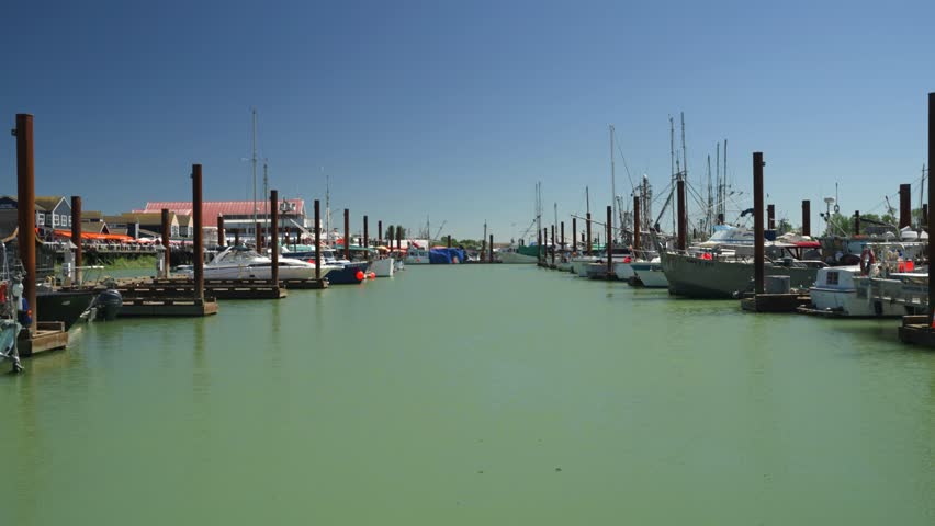 Static handheld shot looking down the marina in a small local fishing village, Steveston, Canada. There are large wooden poles and moored boats on both sides sitting on the sea water on a sunny day
