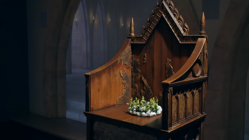 An ornate, weathered wooden throne with intricate carvings stands alone in a dimly lit, arched room. A small floral wreath rests on the seat