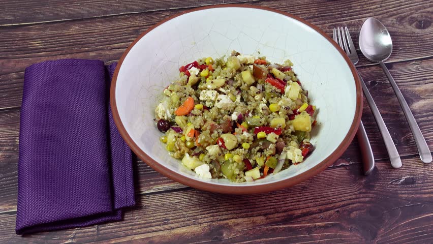 Quinoa salad in a traditional ceramic bowl with spoon, fork and knife and a purple napkin, on a wooden table
