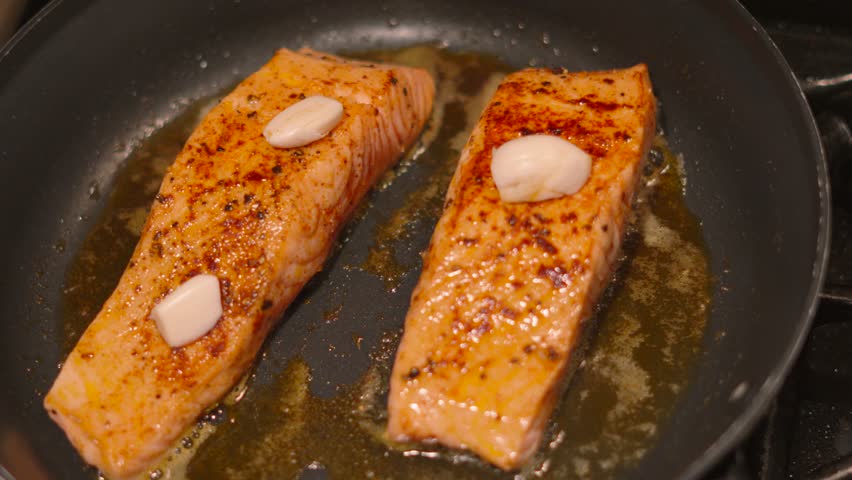 Rotating Overhead View of Wild Caught Salmon Fish Fillets Being Cooked in Frying Pan with Garlic Cloves on Top and Butter Bubbling Underneath. Delicious Healthy Food Footage 4K