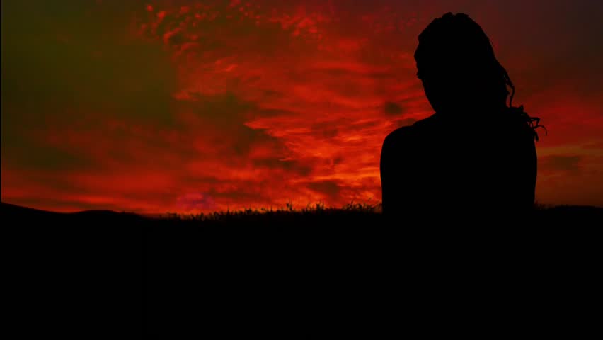 video footage of the silhouette of a woman standing gracefully looking at the twilight view of the red sky with the wind blowing through her hair