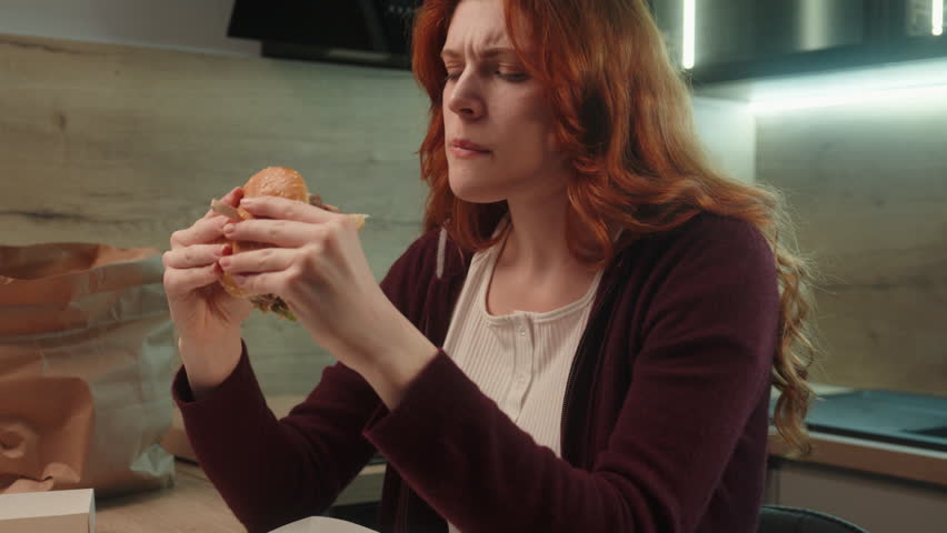 Unhappy displeased Caucasian woman hungry female girl upset lady client customer eating unappetizing cheeseburger chewing disgusting burger taste hamburger junk fast food quality order delivery indoor