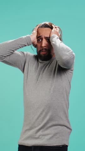 Vertical video Portrait of frightened man covering his face with hands, standing isolated against studio background. Scared person with fearful facial expression suffering shock, feeling distraught