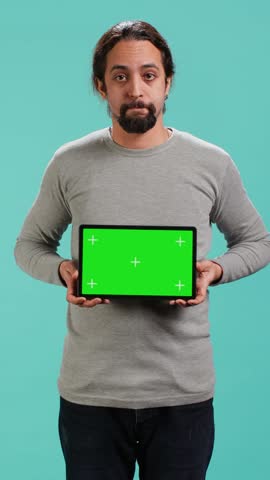Vertical video Influencer presenting green screen tablet, isolated over studio background. Arab person holding blank copy space chroma key device used for advertising brands, studio backdrop, camera A
