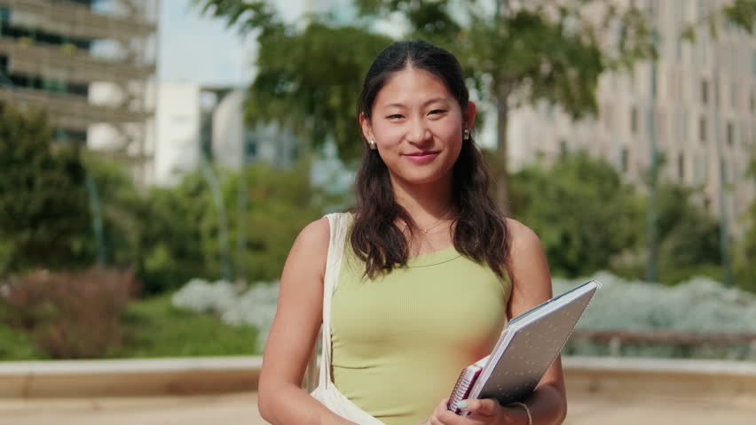 Portrait of a joyful young Asian female student in front of university building on a sunny day. Teenage girl looking at the camera. Education and youth culture concept, milennials
