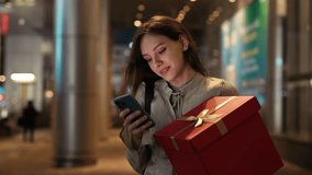 Happy Woman Standing Outside with Big Gift and Chatting in Apps, Smiling to the Screen. Happy Charming Girl in the Street Holding Red Present and Using Cell Phone. Technology and Holiday Concept - Powered by Shutterstock - Get 15% off with code: PIKWIZARD15