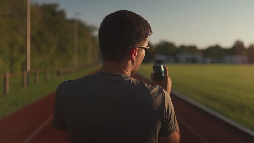 Athletic male massages muscles with hand massage gun, recovering from stadium running workout. . Body treatment with handheld wireless professional vibration shock massager after training session