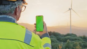 View from behind of mature man in working uniform. Busy engineer or technic with one hand holding smartphone with chroma key. Admiring beautiful sunset with view of large wind turbine. - Powered by Shutterstock - Get 15% off with code: PIKWIZARD15