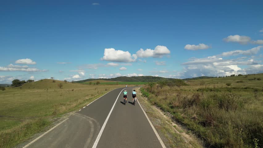 Two cyclists ride down an empty road on a sunny day. The road curves slightly and the cyclists are heading towards a small town in the distance.