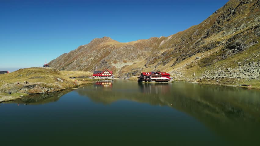 Stunning aerial view of a mountain lake with two charming lodges reflected in the calm water. The surrounding mountains are covered in lush vegetation, creating a picturesque landscape.