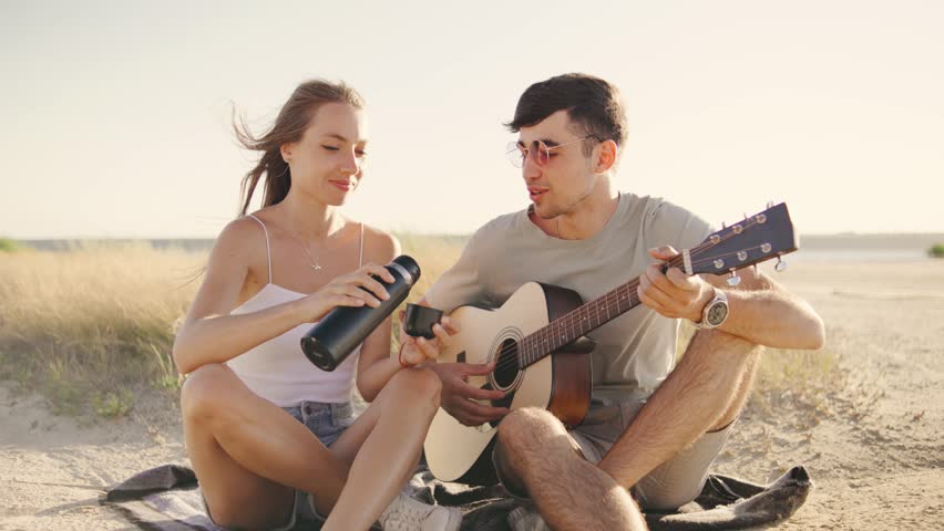 A young man plays guitar and sings a song for his girlfriend as they relax by the lake at sunset, she sipping tea from thermos, enjoying the music and peaceful moment together