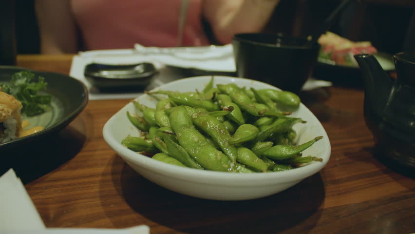A set of Japanese cuisine dishes on a table with focus on a bowl of garlic edamame. Close up shot of plate with boiled soybeans in the center of table with Japanese dishes.