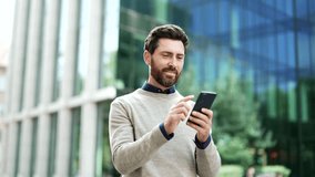 Happy businessman is using a phone standing on the street near a business office building. A smiling handsome bearded male texting, chatting online, browsing web, reads writes messages on a smartphone - Powered by Shutterstock - Get 15% off with code: PIKWIZARD15