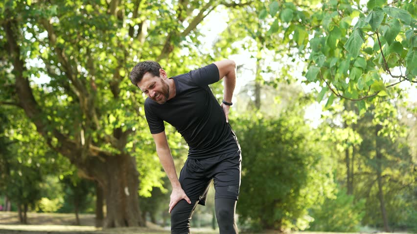 Sportsman suffering from back pain, sports injury, muscle spasm during a morning run in the urban city park. Adult bearded runner having back flank ache and problem after exercise outside in nature