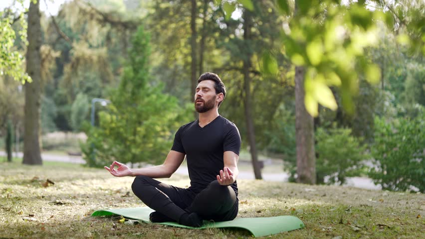 Adult man meditating outdoors in peaceful park environment. Seated on yoga mat amidst trees, focusing on calmness and mindfulness. Concentrated male practices meditation barefoot with his eyes closed