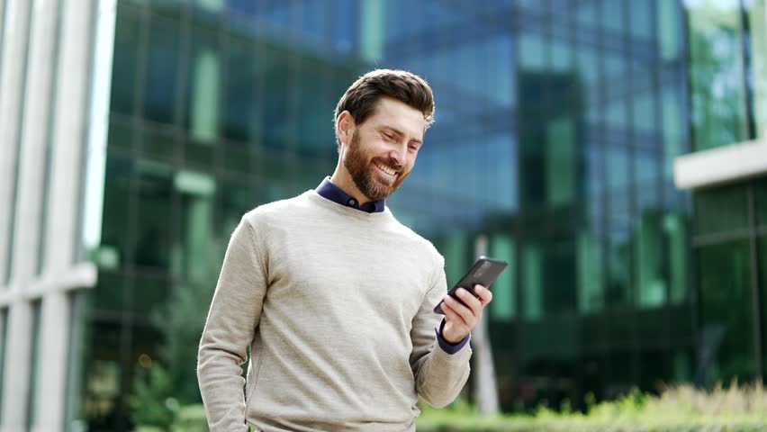 Businessman walking outdoors holding smartphone, smiling while browsing or chatting online. Male professional in casual enjoying city life, checking messages on street near business office building
