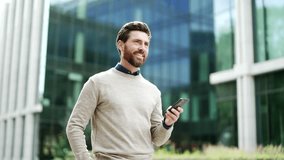 Businessman walking outdoors holding smartphone, smiling while browsing or chatting online. Male professional in casual enjoying city life, checking messages on street near business office building - Powered by Shutterstock - Get 15% off with code: PIKWIZARD15