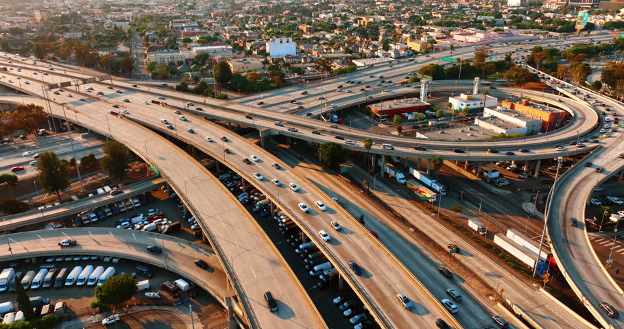 Multiple cars run by the freeways on the interchange and stand parked under it. Lively traffic in Los Angeles, California, the USA at sunset. Top view.