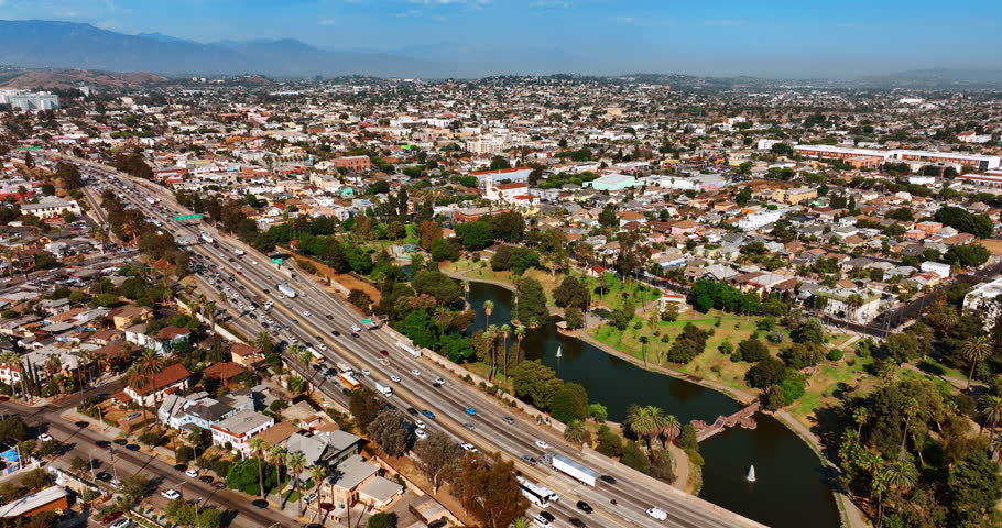 Traffic jam on the wide multi-lane highway in the modern city. Beautiful green park with pond and fountain is nearby the road. Sunny panorama of Los Angeles, California, the USA. Aerial view.