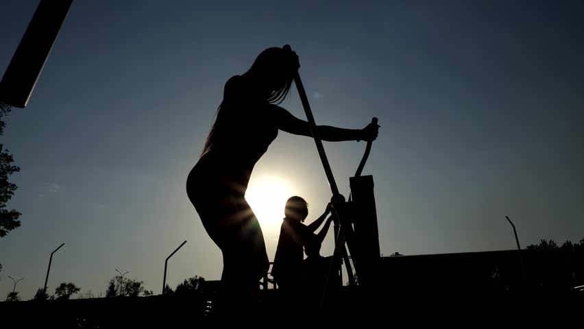 The silhouette of a sporty, pumped-up girl on an orbitrek on an outdoor simulator in the park, the summer setting sun. A sporty woman with a toned body trains on an outdoor sports field. Slow motion.