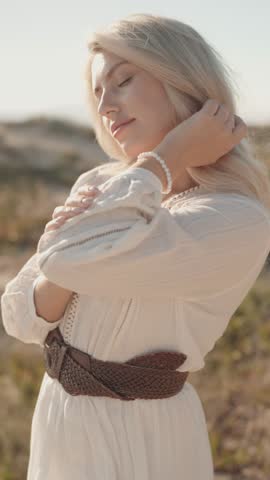 A young, beautiful blonde woman with blue eyes and wearing a white boho dress poses at the beach. She is happy and enjoying life.